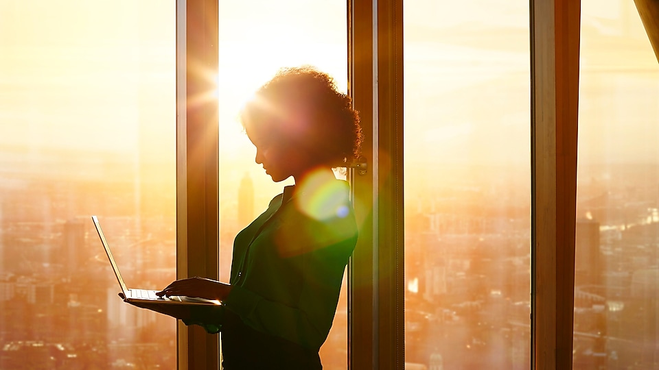 Egy laptop előtt álló nő egy épület ablaka mögött, a lemenő nap fényében