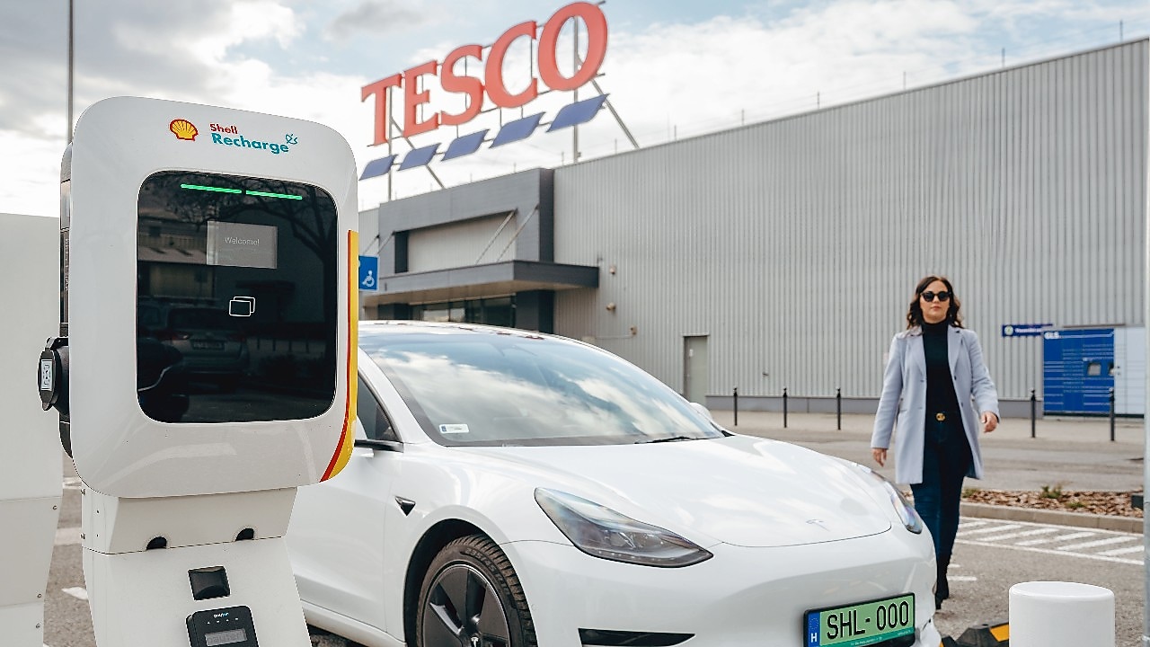 Shell Recharge töltő közelről, a háttérben egy elektromos autóval