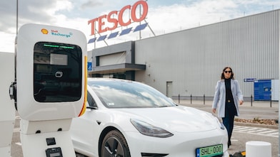 Shell Recharge töltő közelről, a háttérben egy elektromos autóval