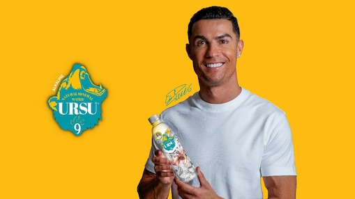 Cristiano Ronaldo mosolyogva tart egy Ursu9 márkájú, ásványvíz palackot egy sárga háttér előtt, bal oldalán a márka logójával.