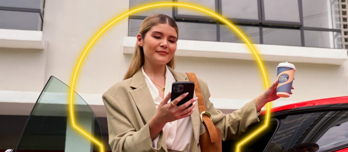 Shelles fogyasztó áll egy piros autó mellett, kezében egy telefonnal és egy kávéval.