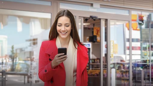 Fiatal nő mosolyogva nézi a telefonját, miközben egy Shell Shop bejáratánál áll