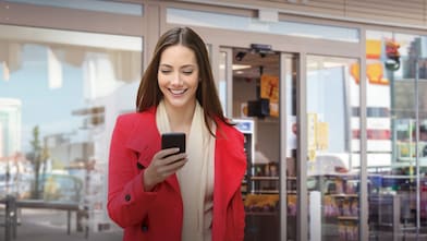 Fiatal nő mosolyogva nézi a telefonját, miközben egy Shell Shop bejáratánál áll