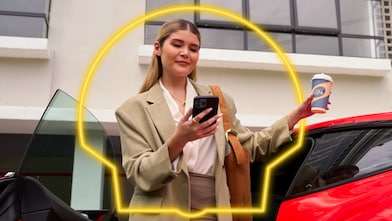 Shelles fogyasztó áll egy piros autó mellett, kezében egy telefonnal és egy kávéval.