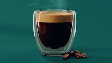 Duplafalú üvegpohárban minőségi espresso kávé, gardag, sötét crema-val a tetején. A pohár mellett pár kávébab látható.