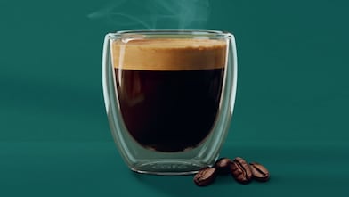 Duplafalú üvegpohárban minőségi espresso kávé, gardag, sötét crema-val a tetején. A pohár mellett pár kávébab látható.